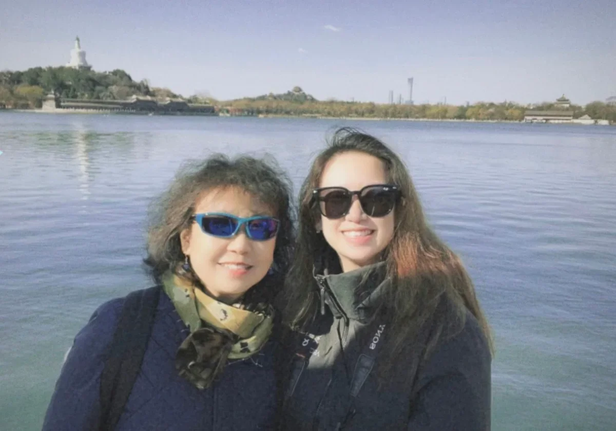 A photo of a mother and daughter in sunglasses and winter coats standing by the lake at Beihai Park, with the White Dagoba on the far shore.