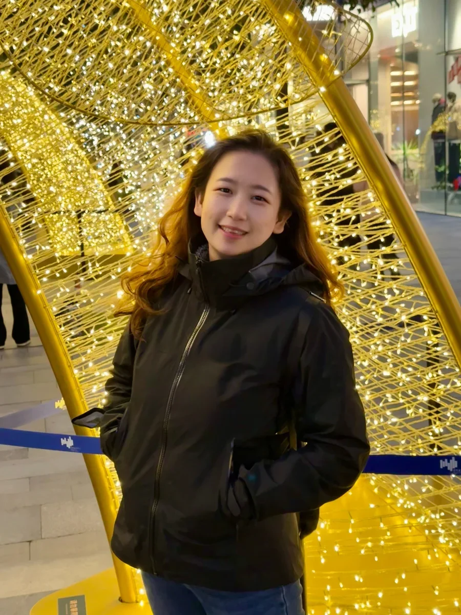 A photo of a smiling woman with long wavy hair in a dark jacket, standing in front of a golden light installation at night.