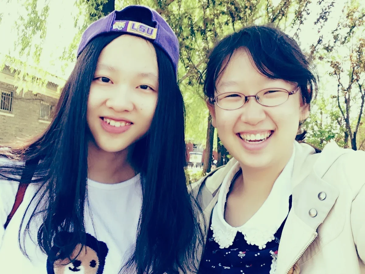 A 2013 outdoor selfie of two smiling young women: one in a purple baseball cap with long dark hair, the other with short dark hair and glasses.