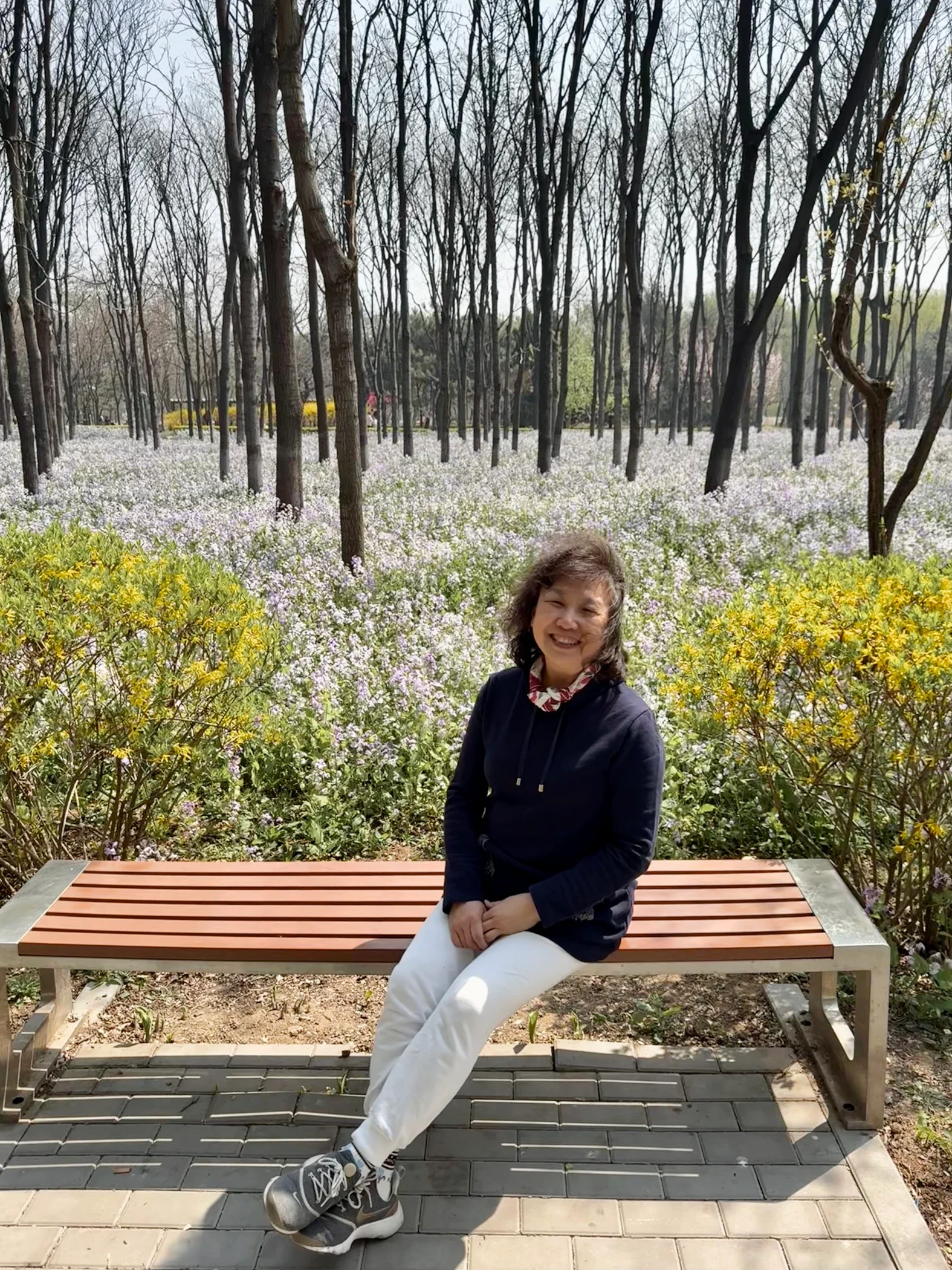 My mom sitting on a wooden park bench among spring flowers.